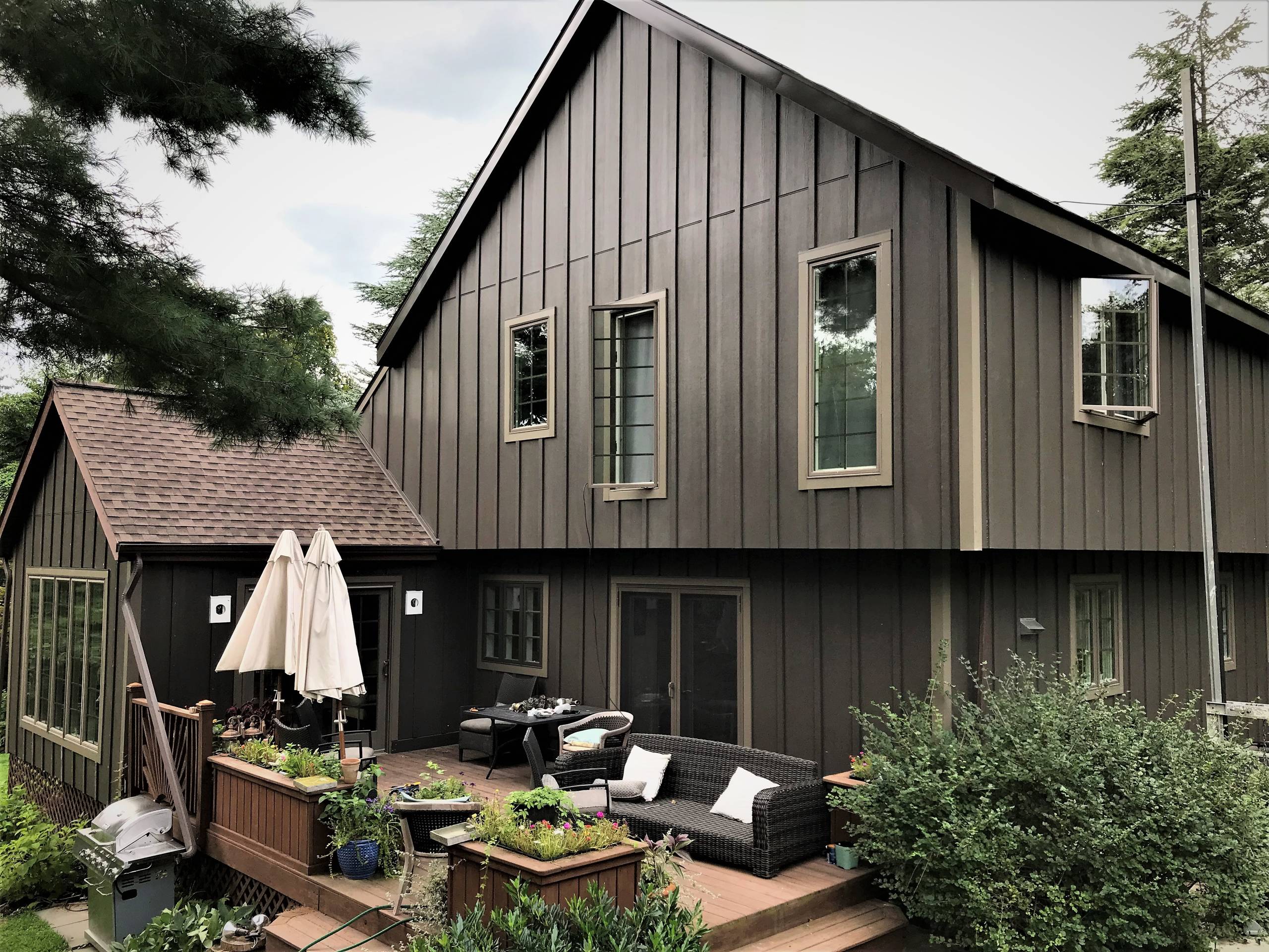 A cozy backyard patio with furniture set in front of a dark brown, two-story house featuring new siding installations & repairs, with trees surrounding the property.