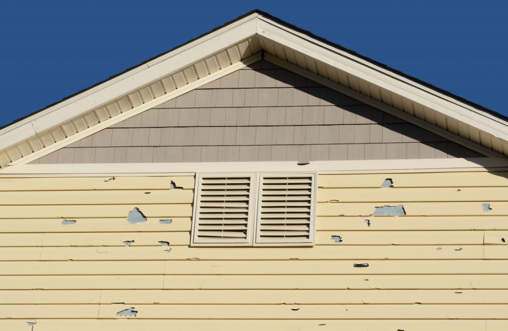 Gable end of a house with peeling yellow paint and a white louvered shutter, in need of siding installations & repairs.